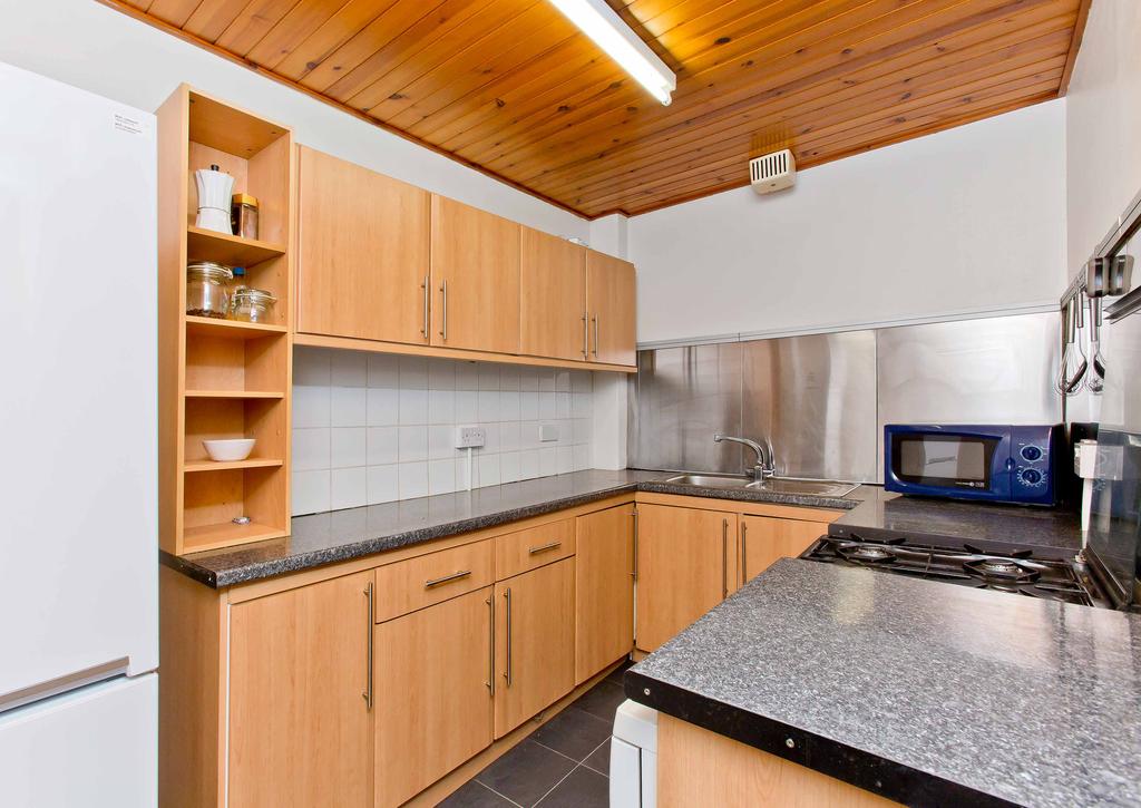 The neighbouring kitchen is fitted with a good selection of modern wood-styled cabinets framed by plentiful workspace, stainless-steel splashbacks, and white wall tiling.